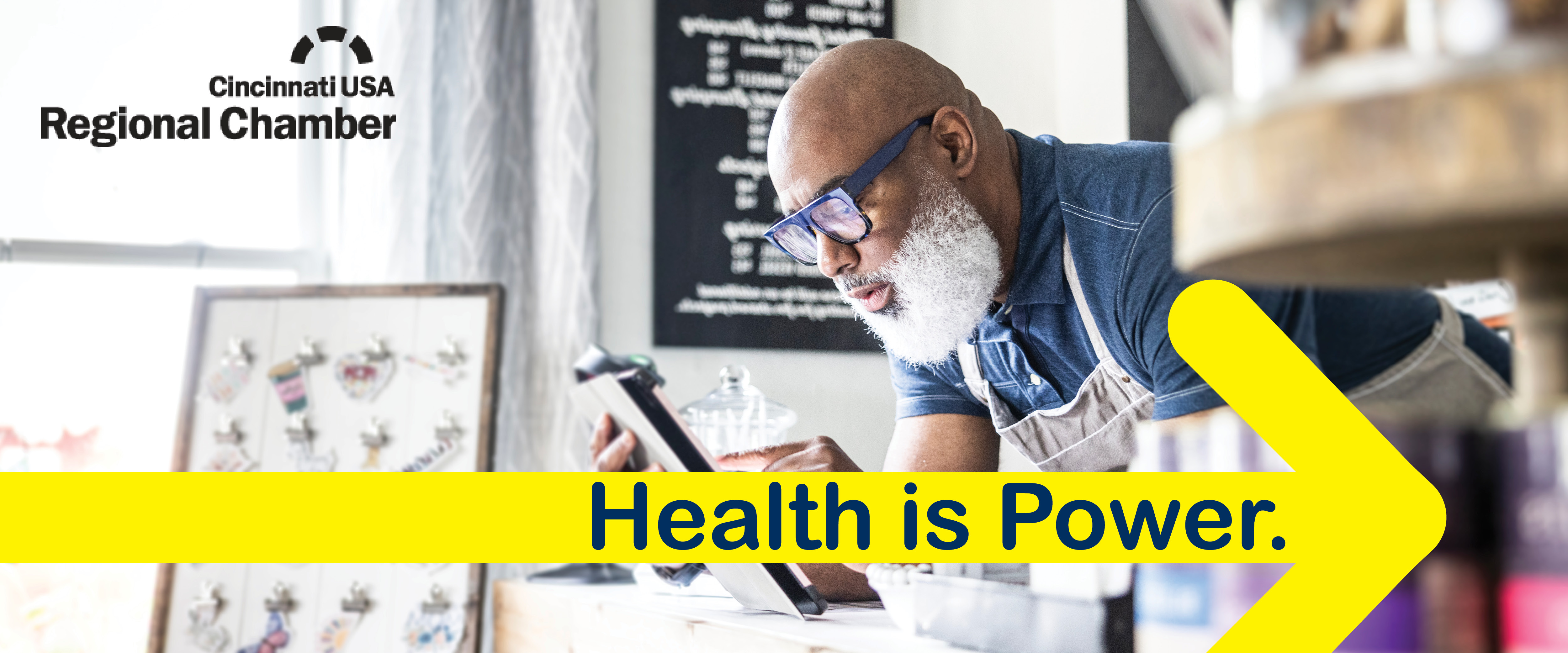 Man working on a tablet at the front desk of a coffee shop with a graphic of an arrow that reads, "Health is Power."