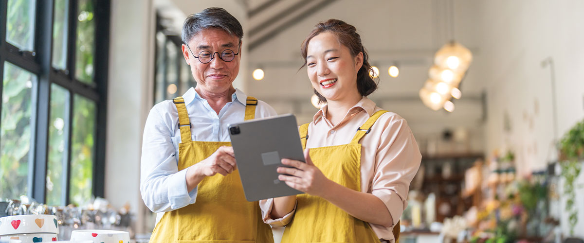 Two small business employees wearing aprons review information on a tablet inside a retail shop or café. The image represents small business management, workplace collaboration, digital tools, employee training, business operations, and access to workplace benefits or business resources.