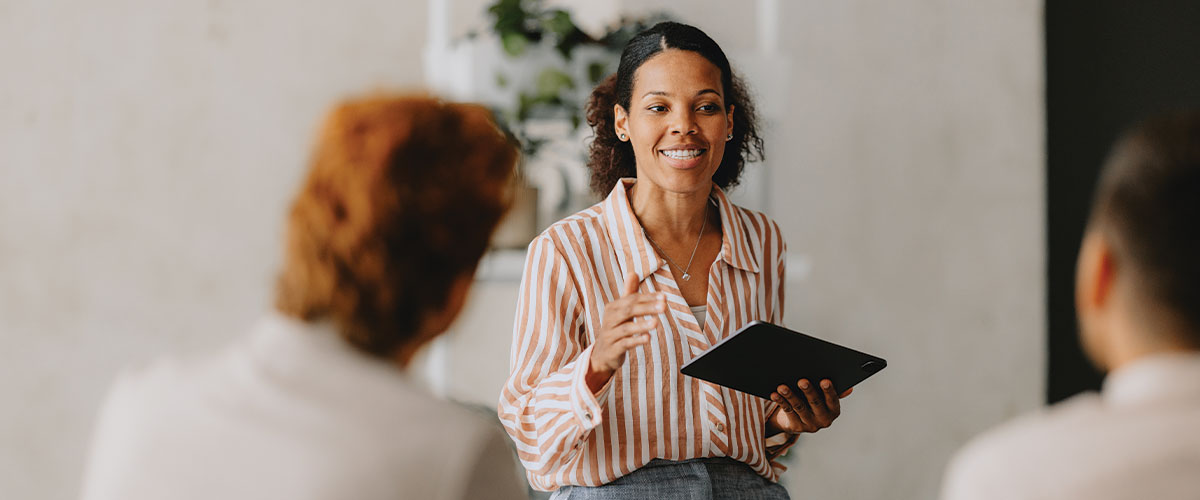 Person presenting information to a small group while holding a tablet during a business meeting or training session. The image represents professional development, broker resources, employee education, workplace collaboration, benefits communication, and business training programs.