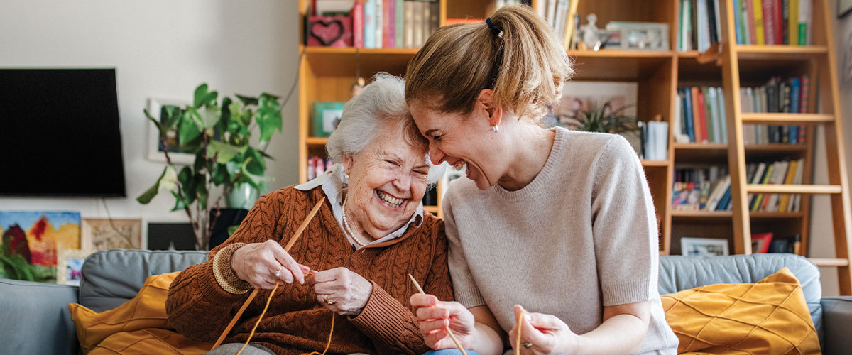 Older adult and caregiver sitting together on a couch while knitting in a home setting, representing Medicare support, healthy aging, long-term care planning, caregiving resources, senior wellness, and maintaining quality of life at home.