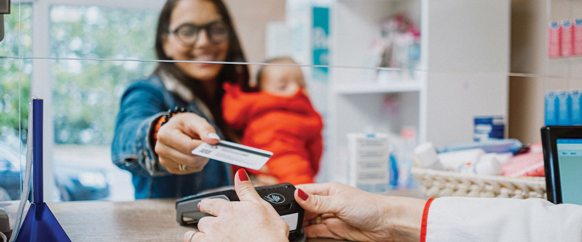 Person presenting a benefits or insurance card at a healthcare or pharmacy service counter while holding a child. The image represents healthcare coverage, flexible spending accounts, health savings accounts, medical benefits, prescription services, and access to healthcare resources for families.