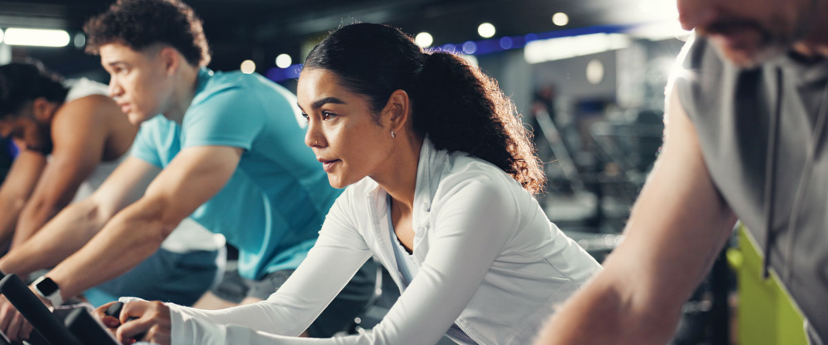 Group fitness participants riding stationary bikes during an indoor cycling class at a gym. The image represents employee wellness programs, physical fitness, preventive health, healthy lifestyles, workplace well-being initiatives, and active health management.