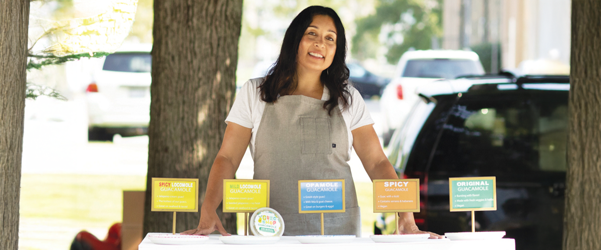 Owner of "The Guac Shop" standing alongside her products at an outdoor event.