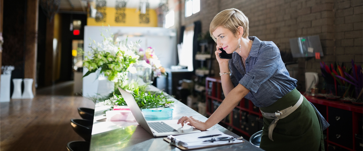 A florist works at a wooden counter, talking on a phone while using a laptop, surrounded by fresh flowers and plants in a bright, modern flower shop.